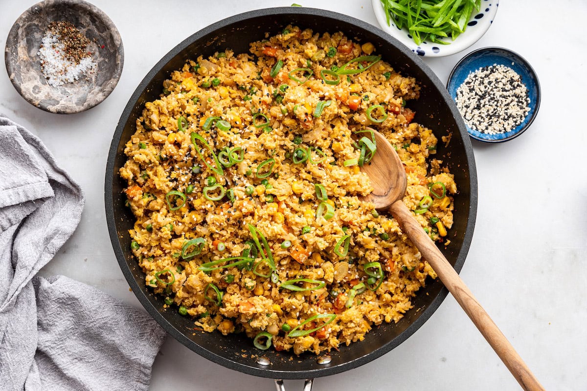 Cauliflower fried rice with eggs in a skillet with a wooden serving spoon. The dish is garnished with fresh green onion.