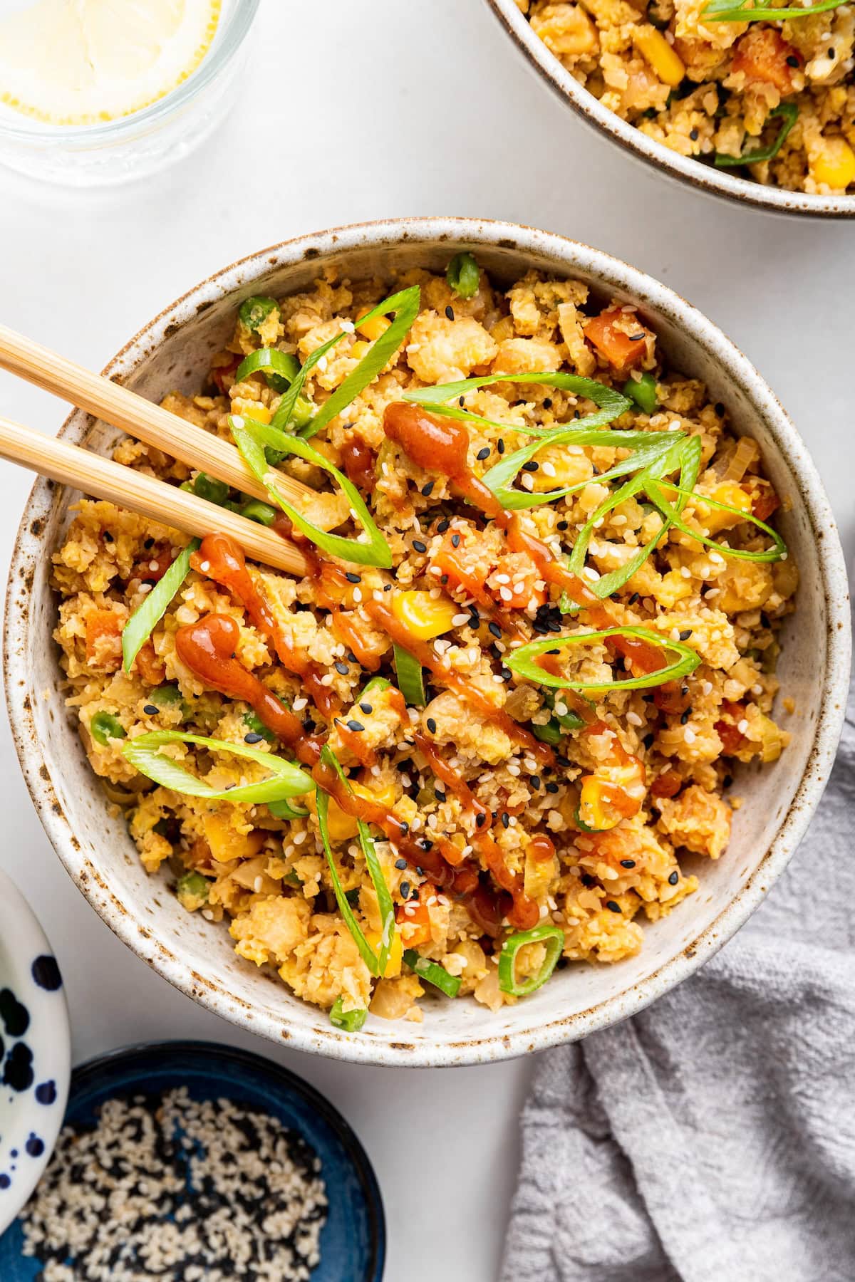 Cauliflower fried rice with egg in a bowl, garnished with fresh green onion and a drizzle of siracha.
