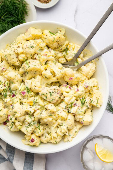Cauliflower potato salad in a large serving bowl with silver serving utensils. The salad is topped with fresh dill.