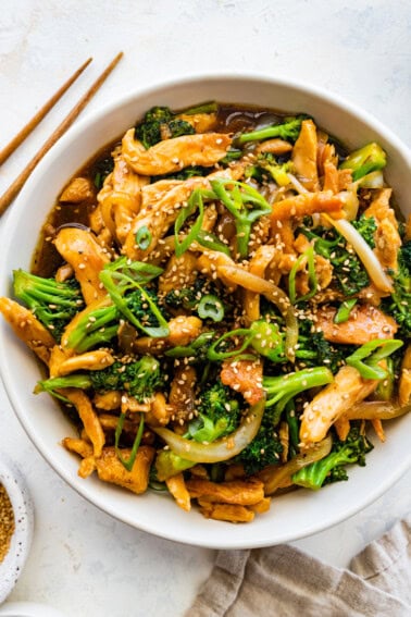 Chicken and broccoli stir fry in a white bowl, garnished with sesame seeds and green onions, with wooden chopsticks beside it.