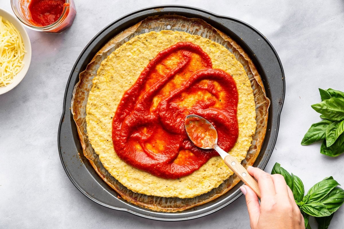 A hand spreading pizza sauce over baked chicken crust with a spoon.