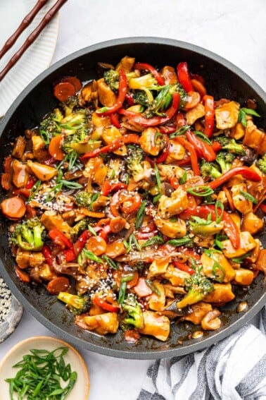 Chicken stir fry with colorful vegetables and tender chicken in a skillet, topped with sesame seeds and green onions.