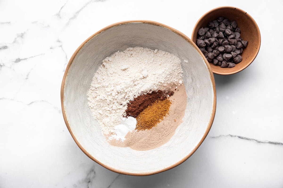 Bowl with oat flour, cocoa powder, protein powder, baking powder, and coconut sugar on a marble counter.