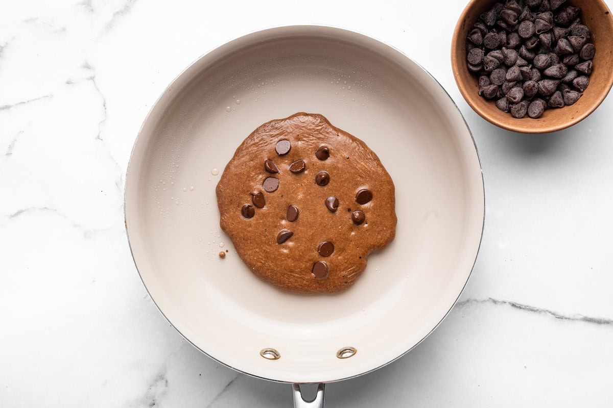 Chocolate protein pancake cooking in a skillet with chocolate chips on top.