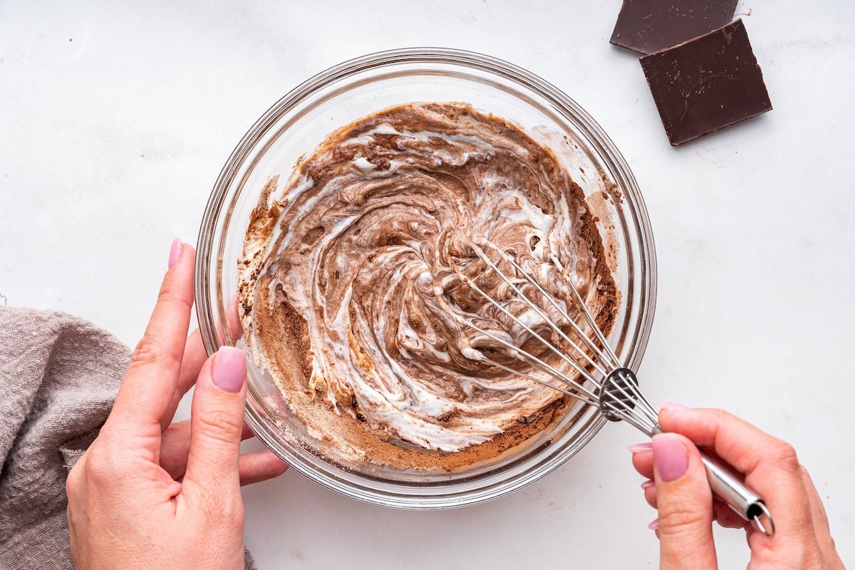 A womans hand uses a metal whisk to combine ingredients for a chocolate protein pudding in a large glass bowl.