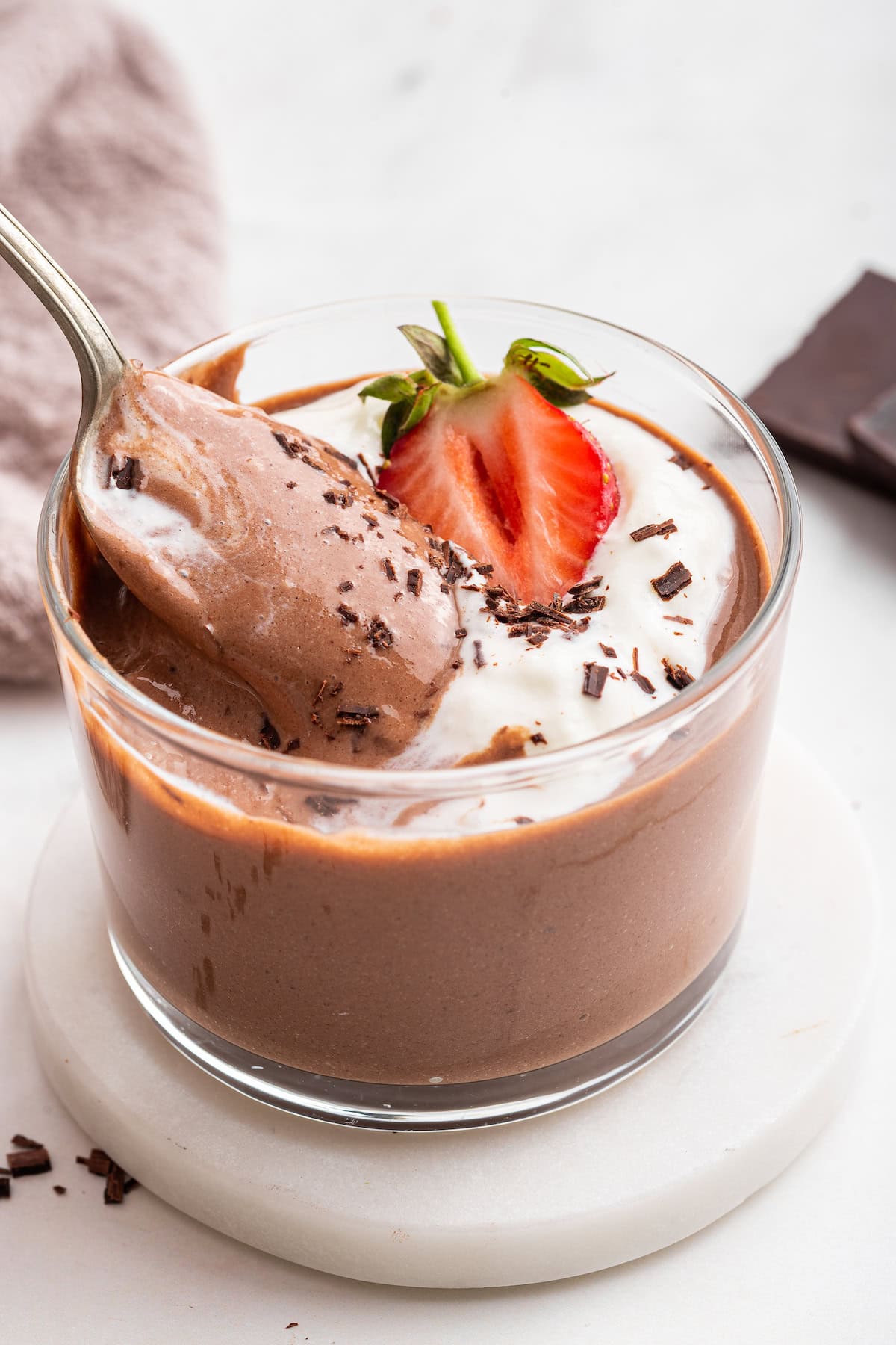 A spoon in a glass cup of chocolate protein pudding that's topped with whipped cream, shaved chocolate, and a strawberry.