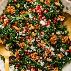 A kale salad topped with fresh pomegranate, goat cheese and candied walnuts. The salad is served in a white bowl with gold salad spoons.