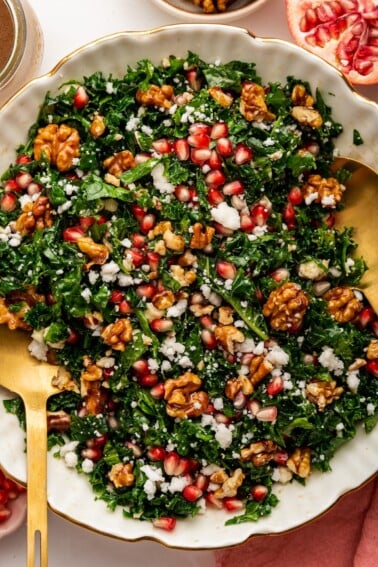 A kale salad topped with fresh pomegranate, goat cheese and candied walnuts. The salad is served in a white bowl with gold salad spoons.