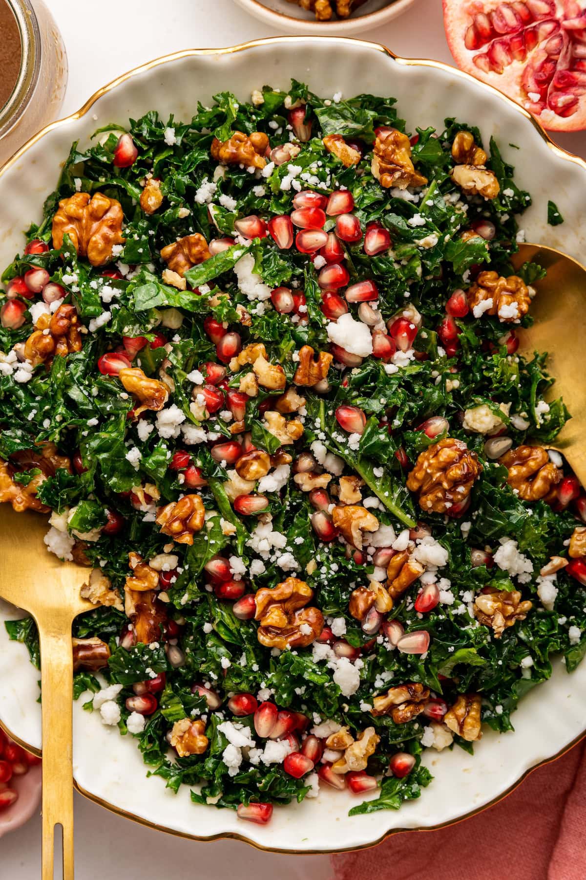 A kale salad topped with fresh pomegranate, goat cheese and candied walnuts. The salad is served in a white bowl with gold salad spoons.