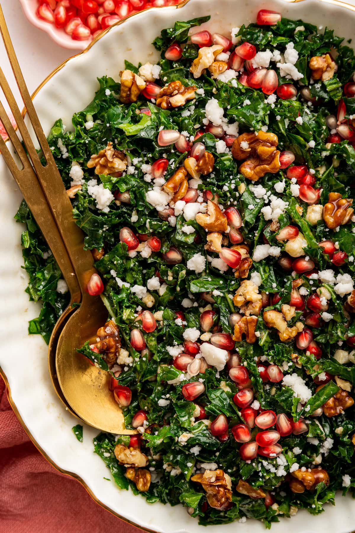 A kale salad topped with fresh pomegranate, goat cheese and candied walnuts. The salad is served in a white bowl with gold salad spoons.