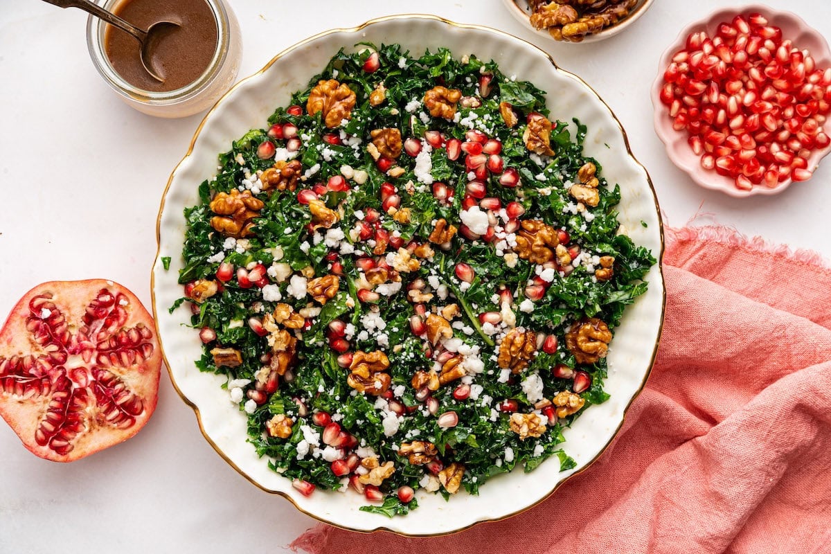 A kale salad topped with fresh pomegranate, goat cheese and candied walnuts. The salad is served in a white bowl.