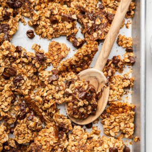 Clusters of chocolate chip cookie granola on a silver baking sheet served with a wooden spoon.