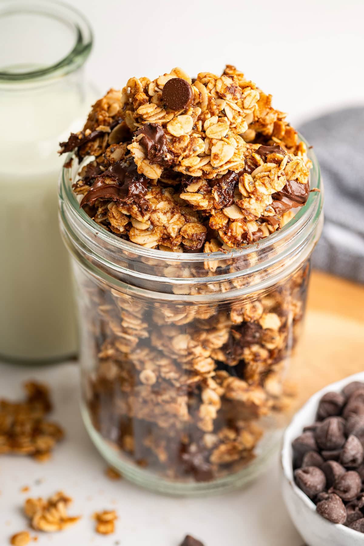 Chocolate chip cookie granola in a glass storage jar. The granola is overflowing from the top of the glass and it's served alongside milk.