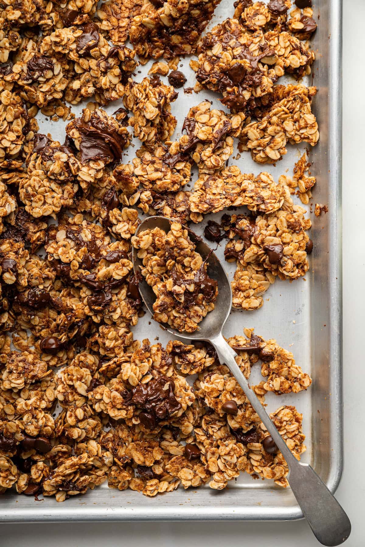 Clusters of chocolate chip cookie granola on a silver baking sheet served with a silver spoon.