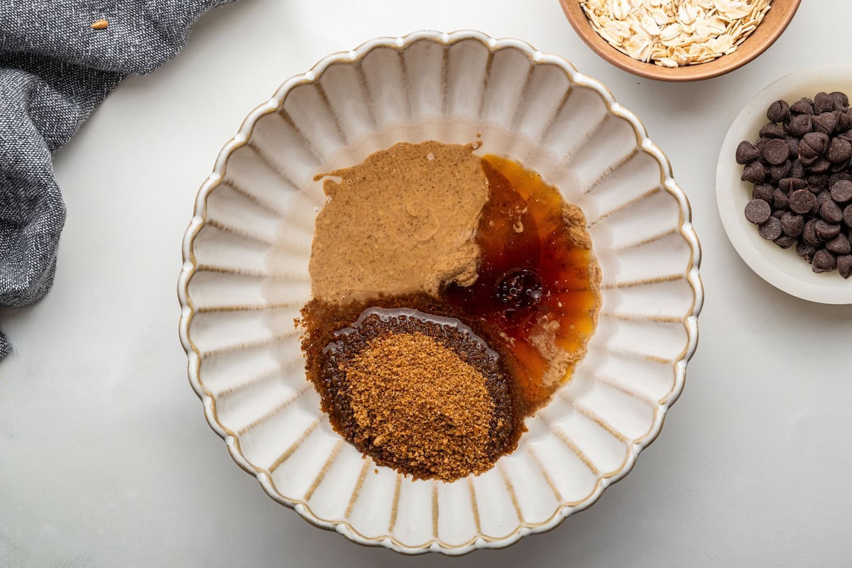 The wet ingredients for the chocolate chip cookie granola in a ceramic mixing bowl: almond butter, coconut oil, maple syrup, coconut sugar and vanilla.