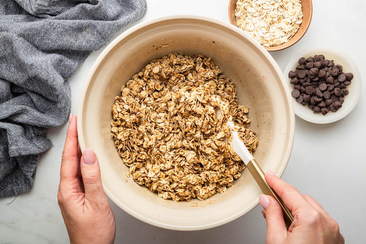 A woman's hands mixing together the cookie granola iwith a white spatula n a ceramic mixing bowl.