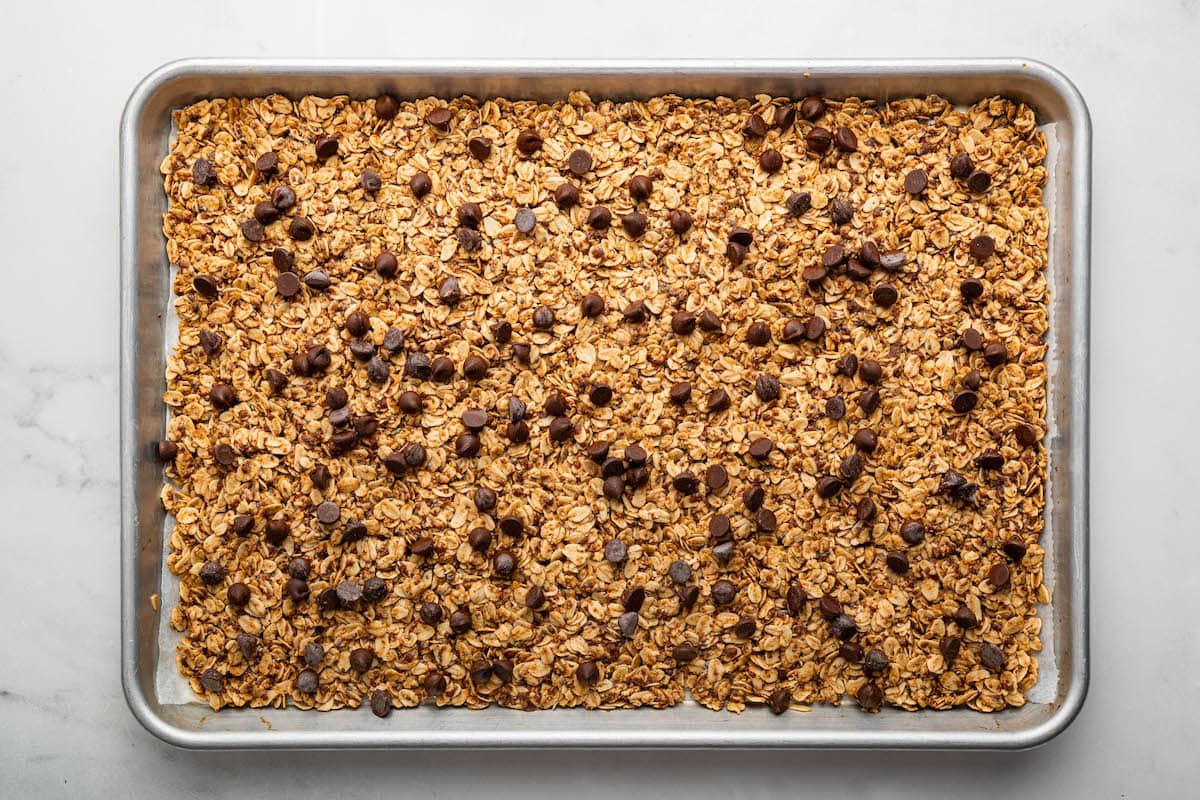 The baked cookie granola topped with chocolate chips spread out on a silver baking sheet lined with parchment paper.