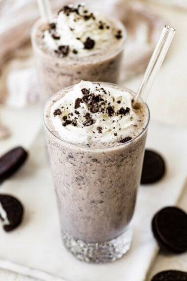 Two cookies and cream protein shakes in glasses, topped with whipped cream and crushed chocolate sandwich cookies, with clear straws inserted.