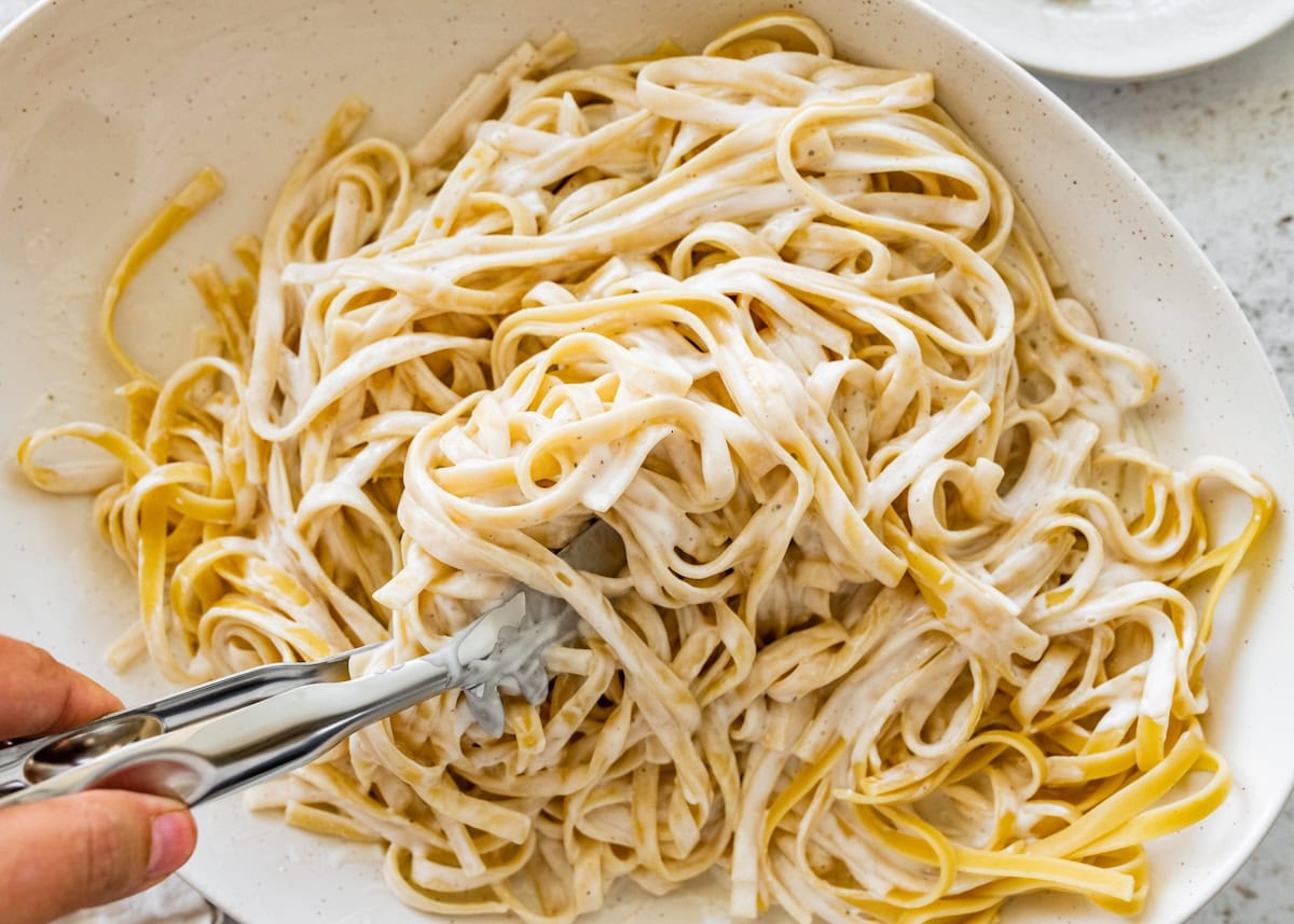 Tongs tossing fettuccine in creamy cottage cheese Alfredo sauce inside a large serving bowl.