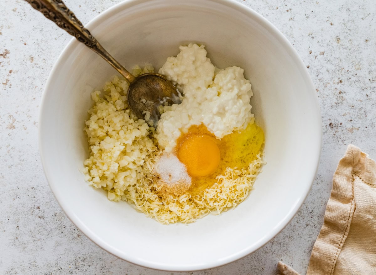 Cottage cheese, cauliflower rice, egg, salt, parmesan cheese, and garlic powder in a mixing bowl with a silver spoon.