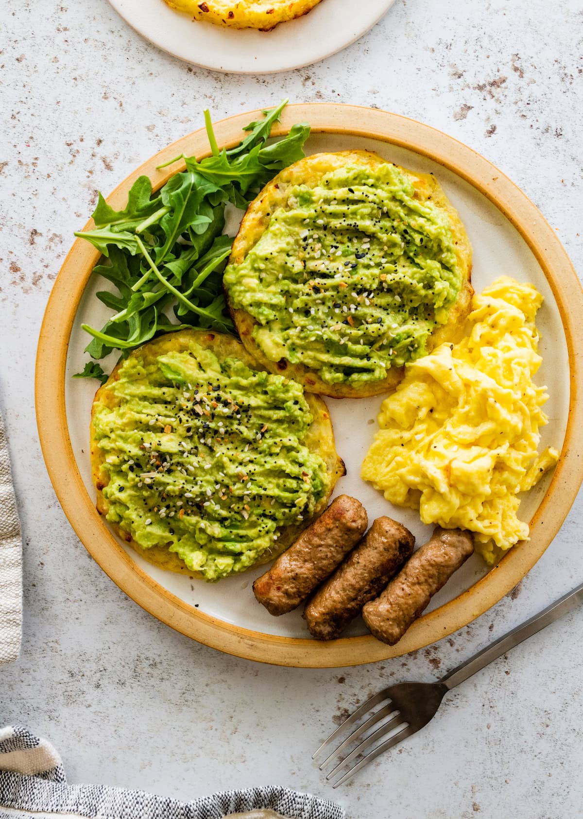 Two cottage cheese cauliflower thins served on a plate with three turkey sausages, arugula and scrambled eggs. The cauliflower thins are topped with mashed avocado and everything seasoning.
