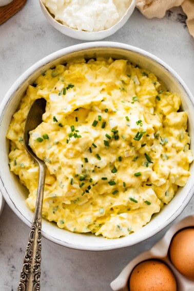 Cottage cheese egg salad served in a white ceramic bowl served with a silver spoon. Fresh chives are topping the egg salad.