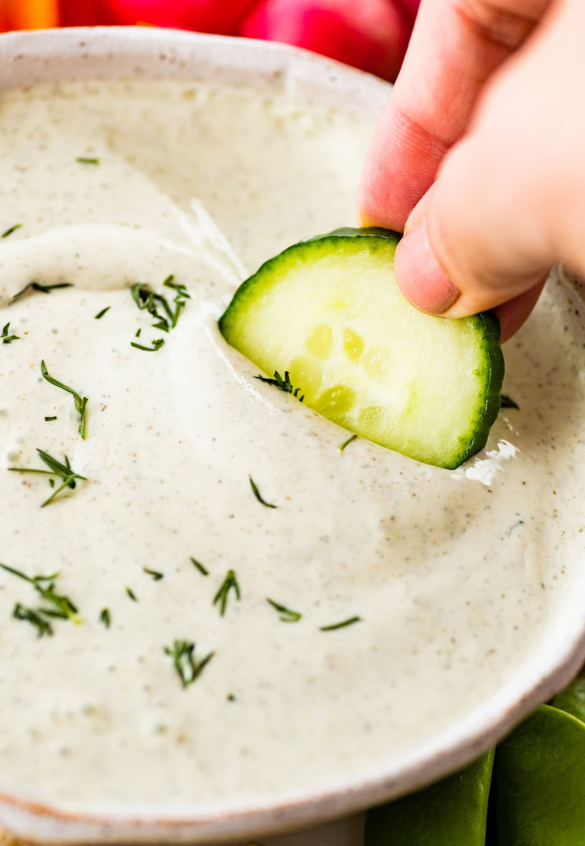 A hand dipping a fresh cucumber slice into a bowl of cottage cheese ranch dip garnished with fresh dill.