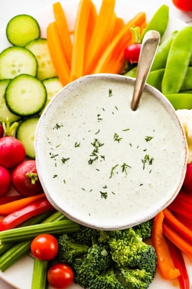 Cottage cheese ranch dip in a white bowl garnished with fresh dill. The bowl is on a plate with an assortment of fresh vegetables.