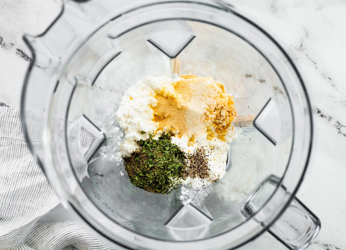 Cottage cheese in a blender with multiple herbs and spices before being blended together for a cottage cheese ranch dip.