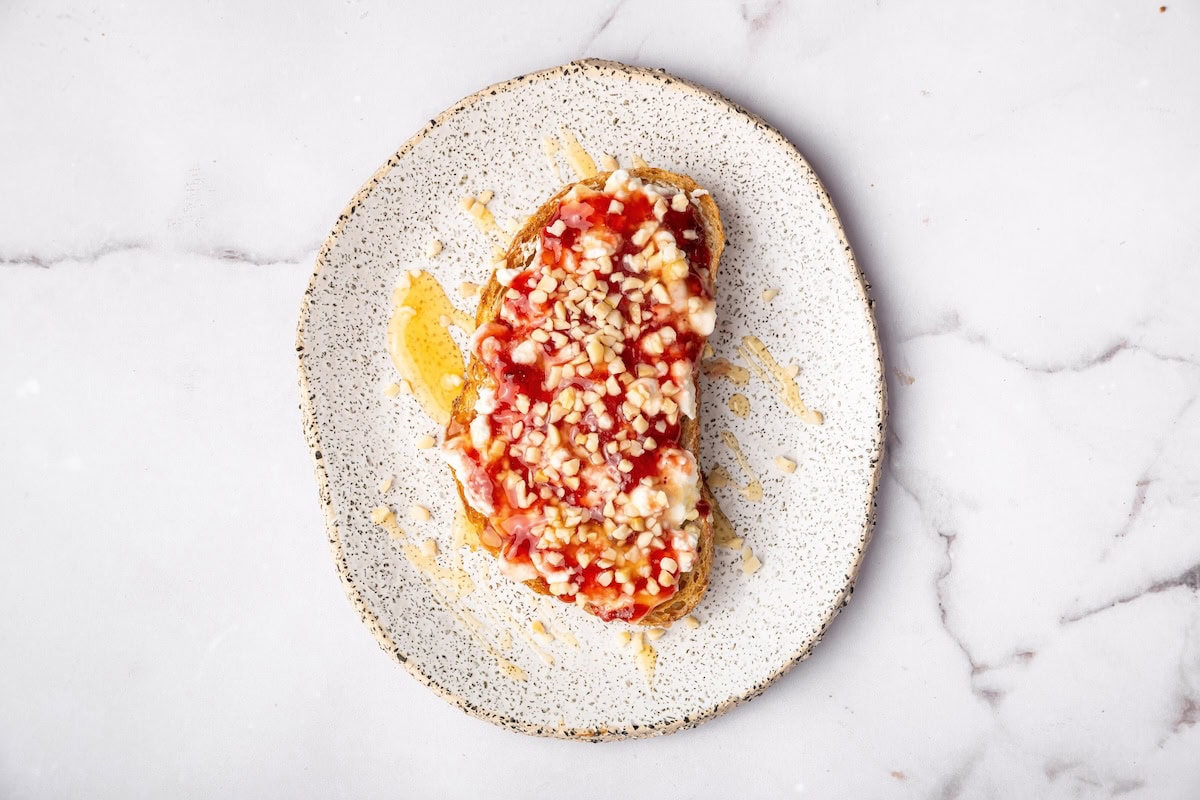 Sweet cottage cheese toast topped with berry jam, chopped nuts, and a drizzle of honey on a speckled plate.