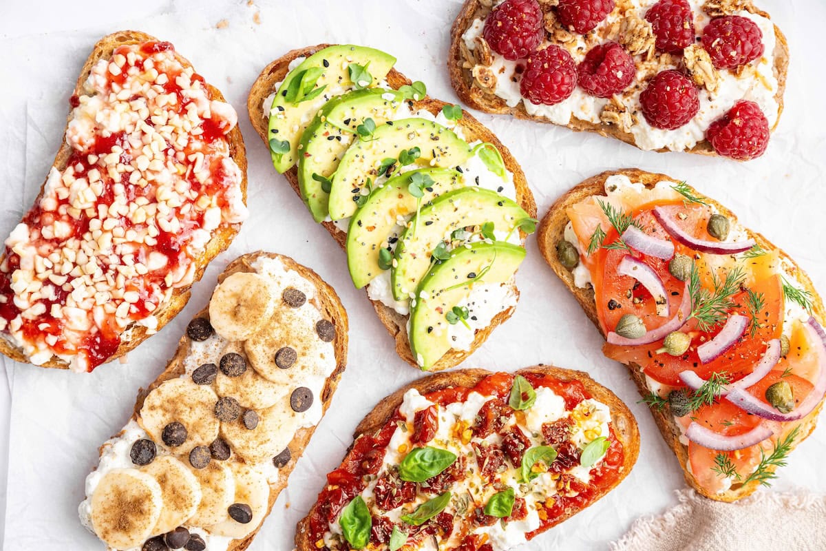Assorted cottage cheese toast ideas topped with avocado, smoked salmon, pizza marinara, raspberries, banana with chocolate chips, and berry jam.