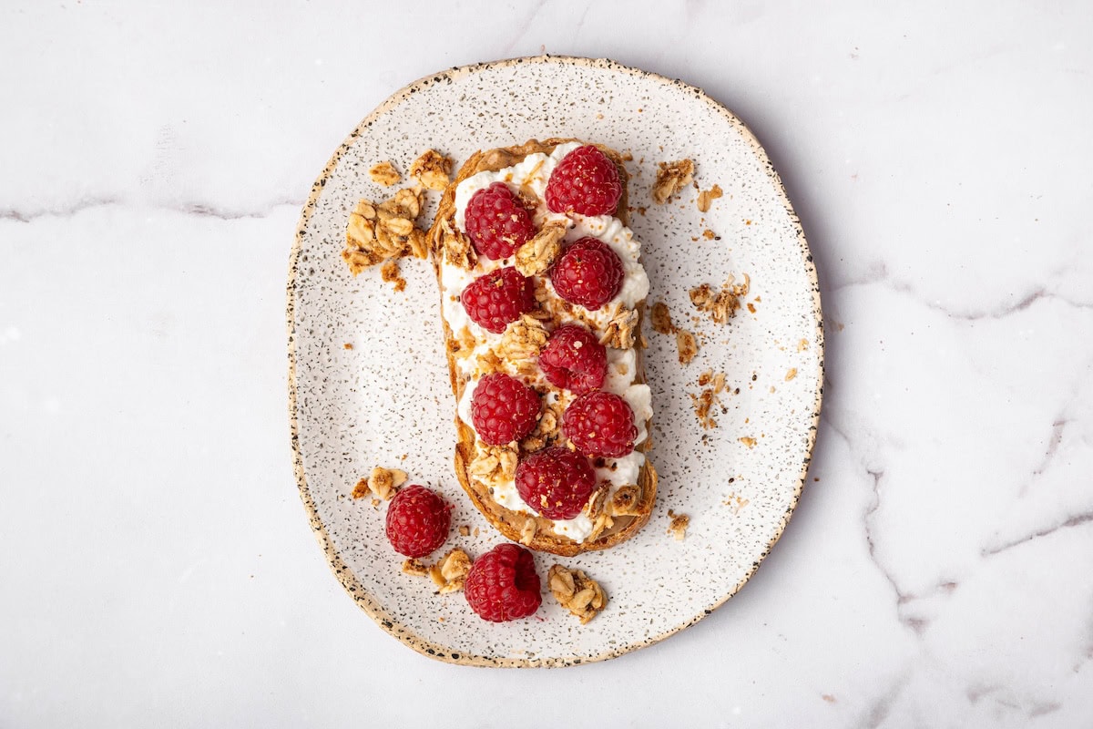 Cottage cheese toast topped with fresh raspberries and crunchy granola on a speckled plate.