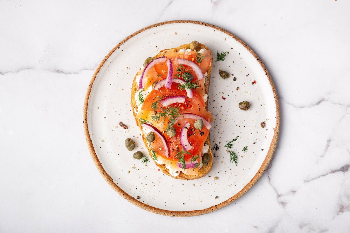 Cottage cheese toast topped with smoked salmon, red onion, fresh dill, and capers on a white plate.