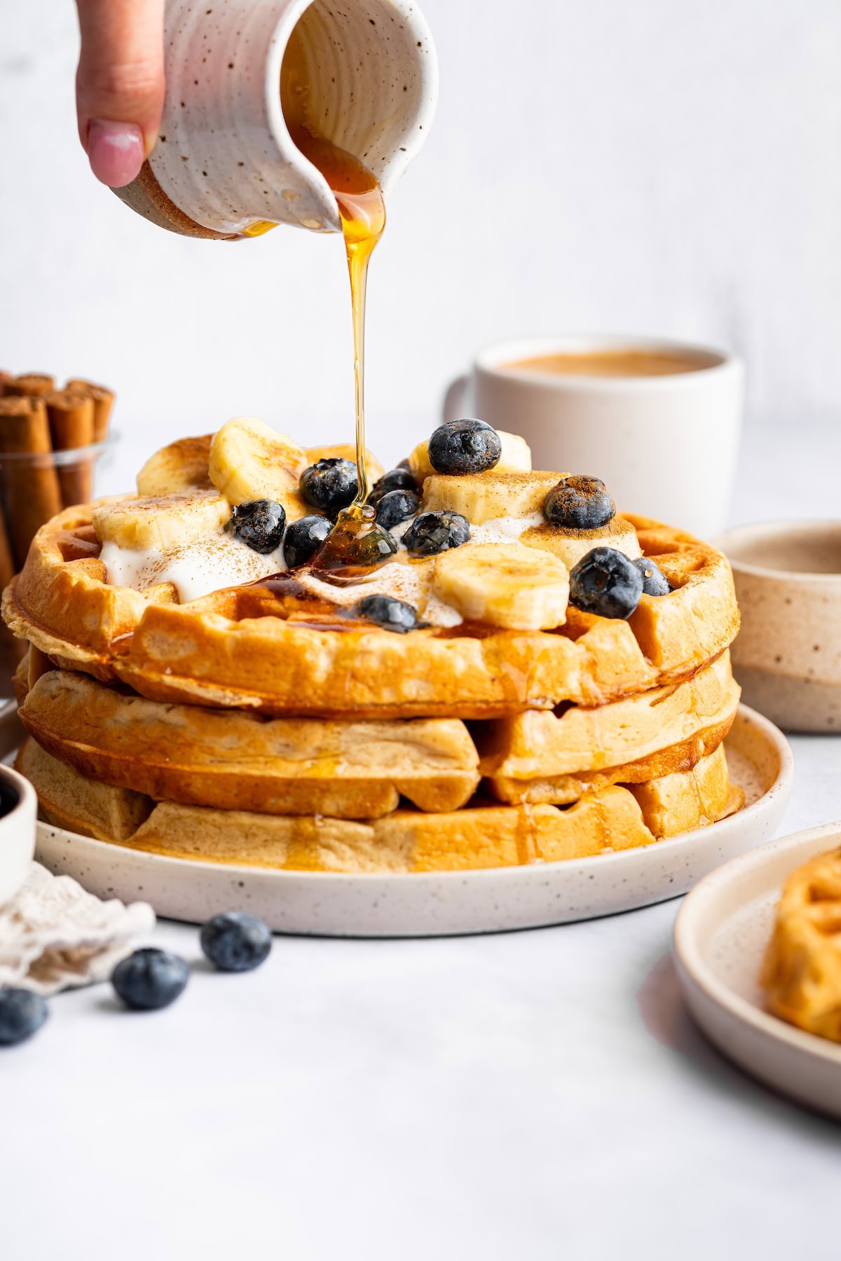 Stack of waffles topped with bananas and blueberries as maple syrup is drizzled over the top.