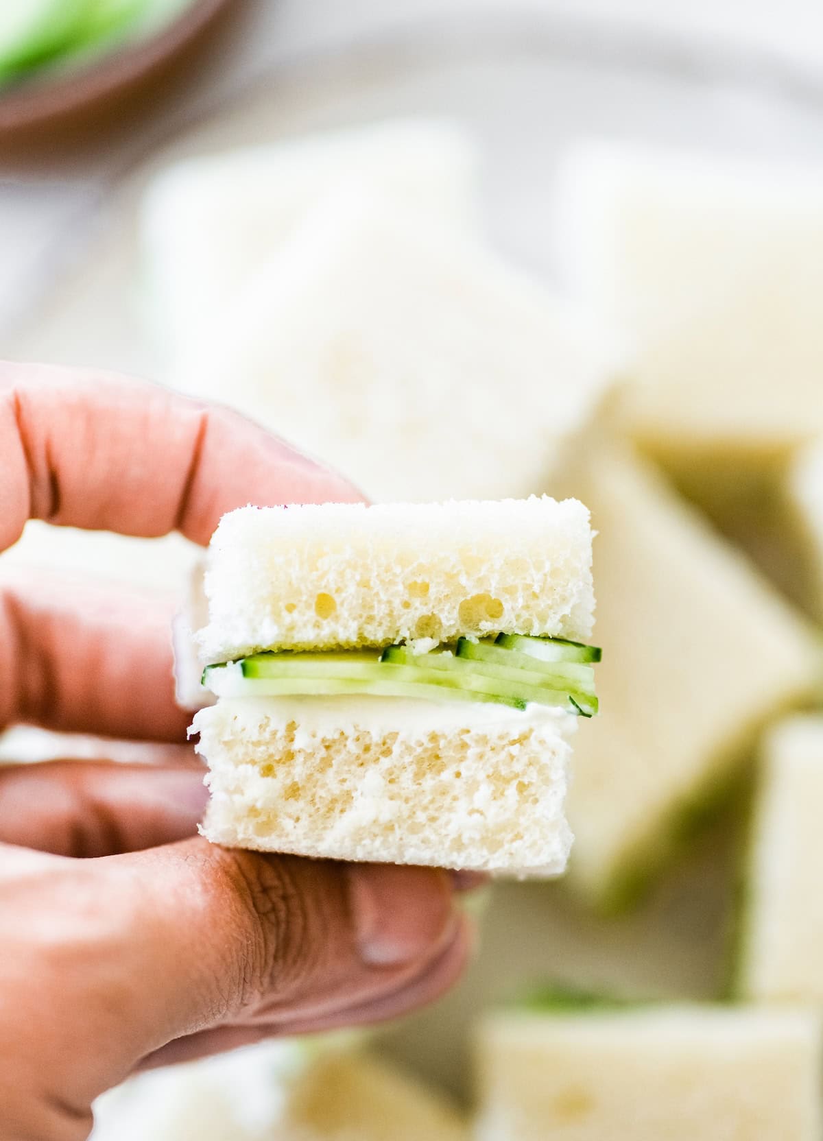 Hand holding a small cucumber sandwich made with soft white bread and thin cucumber slices.