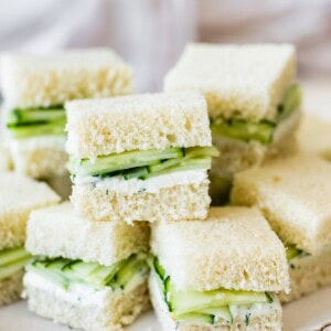 Pile of small cucumber tea sandwiches stacked on a white plate.