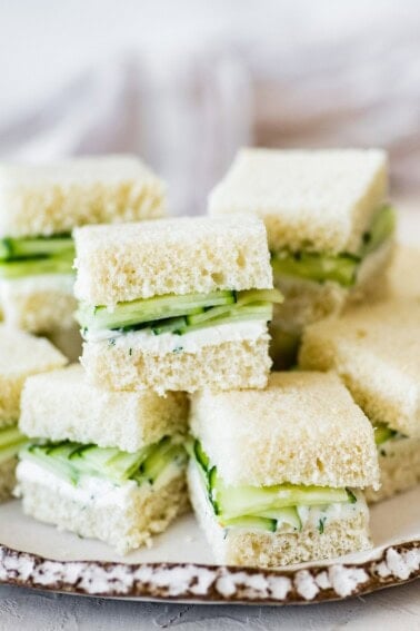 Pile of small cucumber tea sandwiches stacked on a white plate.