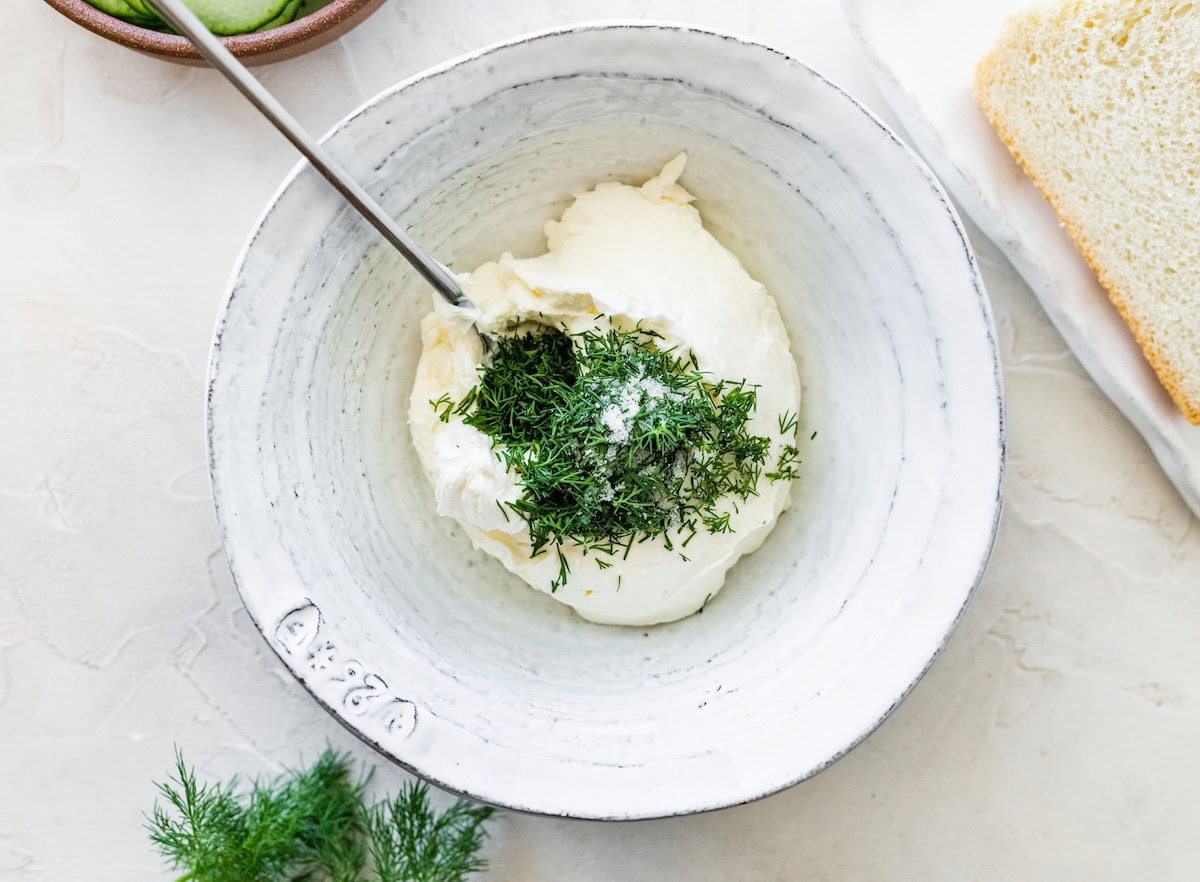Mixing bowl with cream cheese and fresh dill being stirred together with a silver spoon.