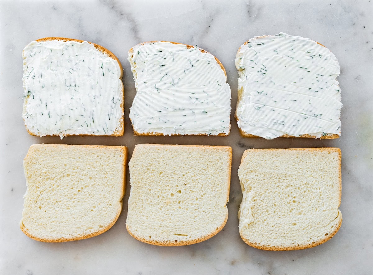 Three bread slices spread with dill cream cheese on a marble countertop.