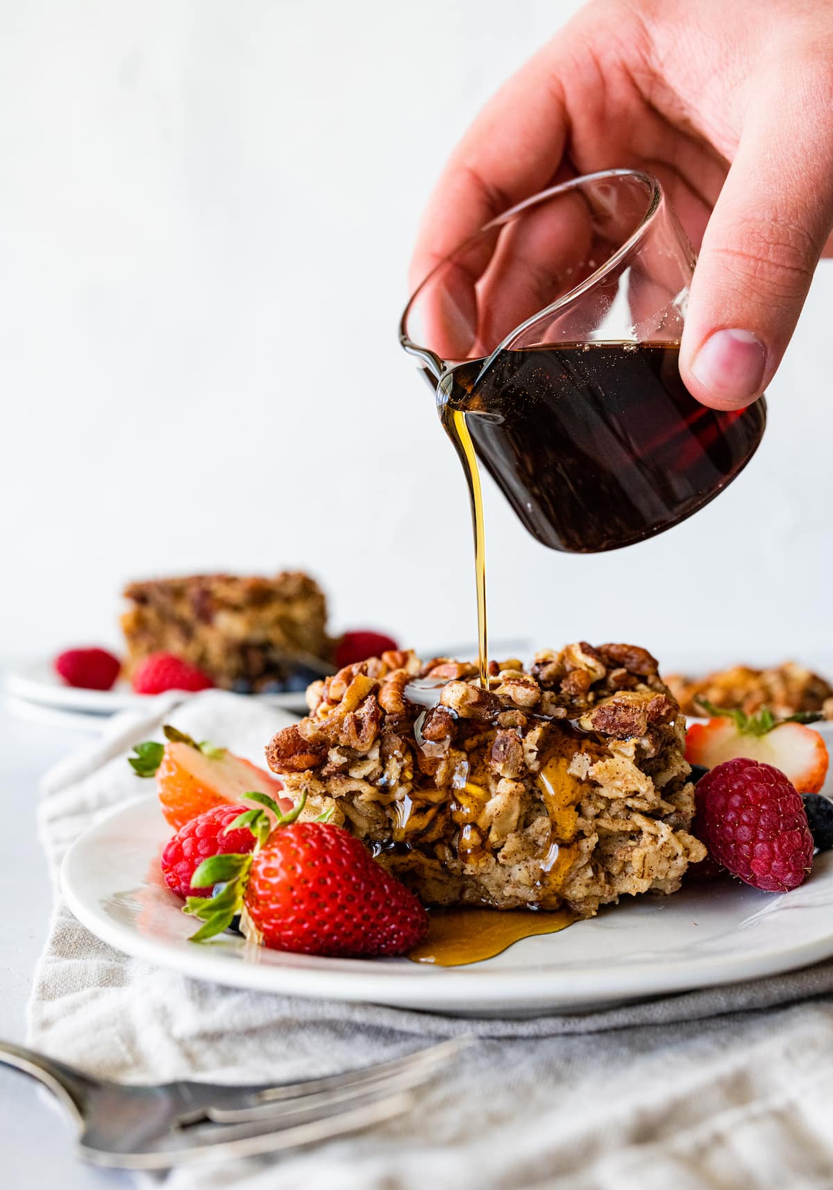 Woman's hand pouring maple syrup over a slice of baked oatmeal on a plate served with fresh strawberries.