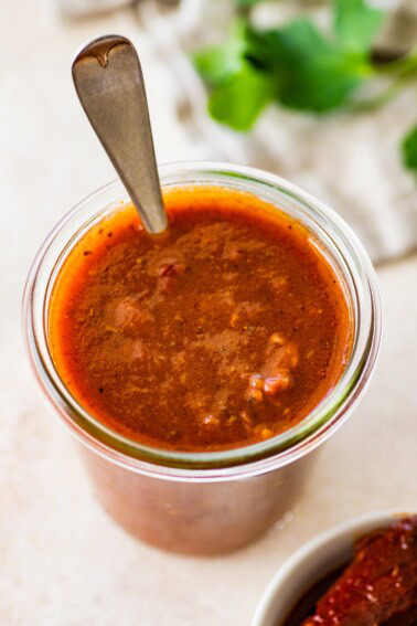 A glass weck jar filled with homemade red enchilada sauce. There is a silver spoon in the glass jar.