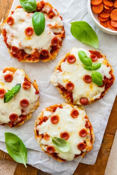 English muffin pizzas topped with melted cheese, mini pepperoni and basil leaves, served on parchment.