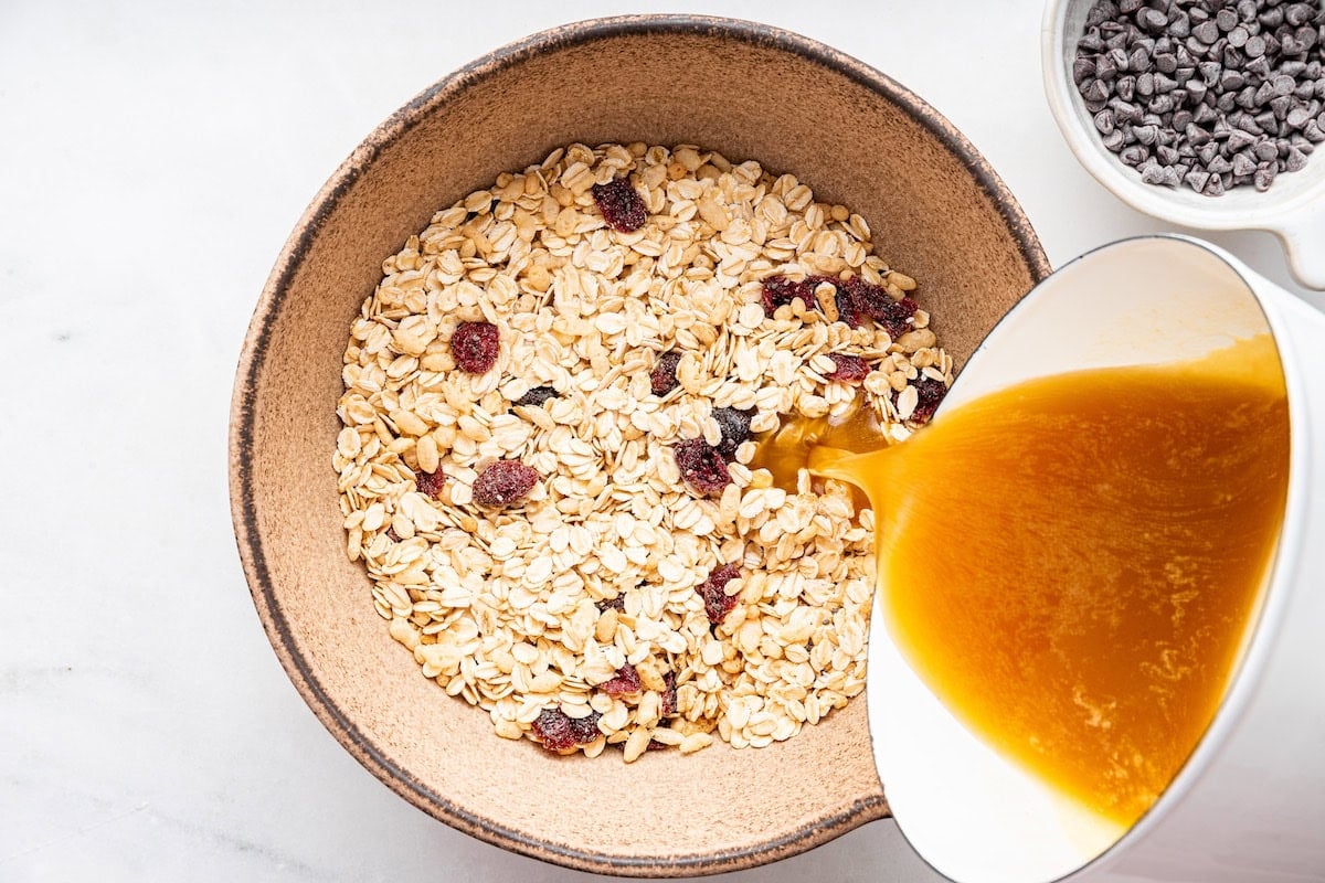 The butter, sugar, and honey mixture is being poured into a bowl of dry ingredients for granola bars.
