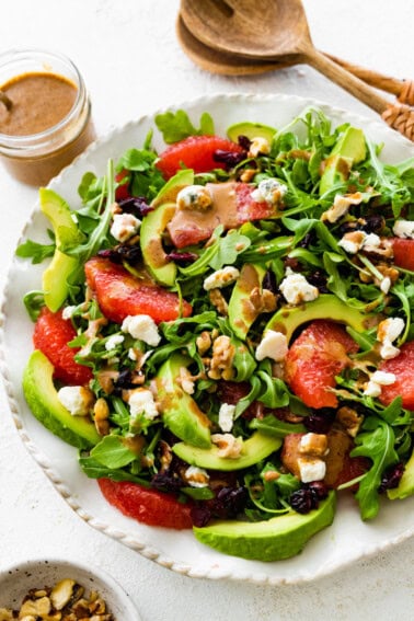 A salad with arugula, avocado, blue cheese, grapefruit, nuts, and cranberries on a white plate.