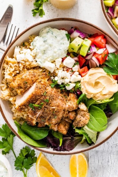 A bowl served with mixed greens, Greek chicken, tzatziki, hummus, cucumber tomato salad and feta cheese.