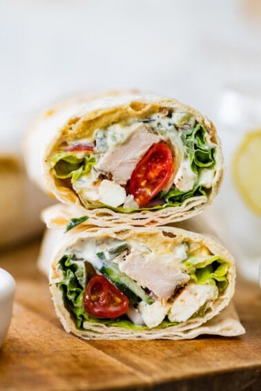 Two halves of a Greek chicken wrap stacked on top of each other on a wooden cutting board. You can see the tomatoes, chicken, lettuce and sauce inside the wrap.