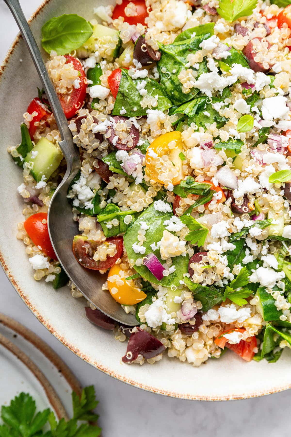 Greek quinoa salad topped with crumbled feta cheese in a tan serving bowl with a silver spoon.