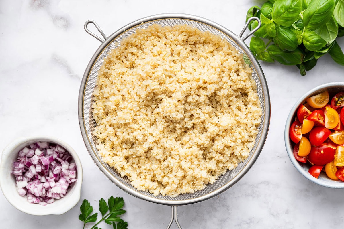 The cooked quinoa in a mesh strainer next to a bowl with cherry tomatoes and a bowl with diced red onion.