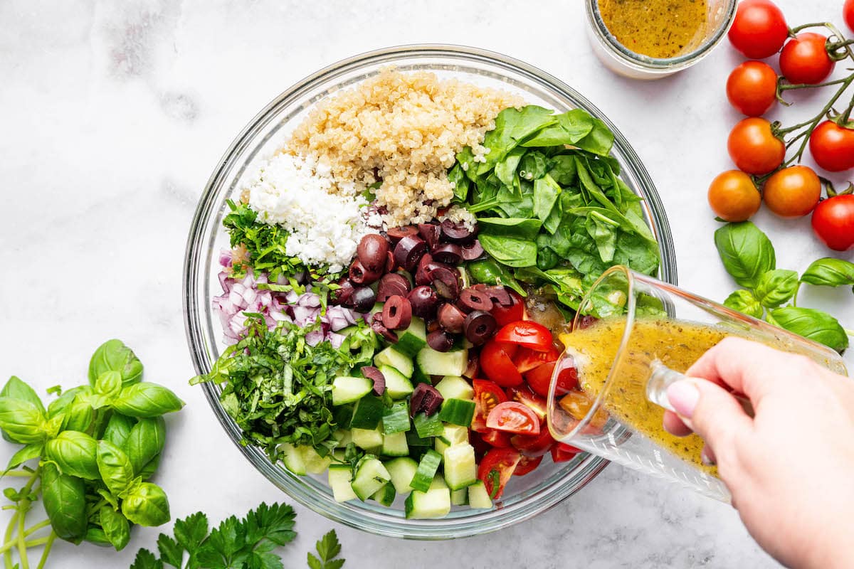 A woman's hand pouring the Greek dressing over the Greek quinoa salad ingredients: cucumber, tomato, kalamata olives, basil, spinach, quinoa, feta and red onion.
