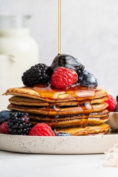 Maple syrup being poured on top of a stack of six greek yogurt pancakes on a plate. They're topped with fresh blueberries, blackberries and raspberries.